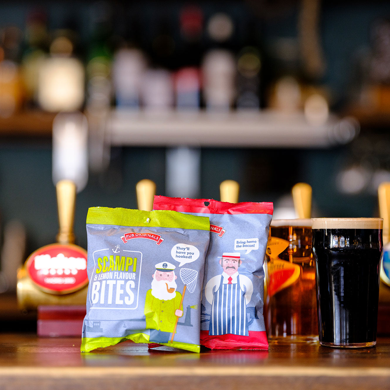 Two packages of Pub Originals snacks on a bar counter with two beers and a beer tap in the background.
