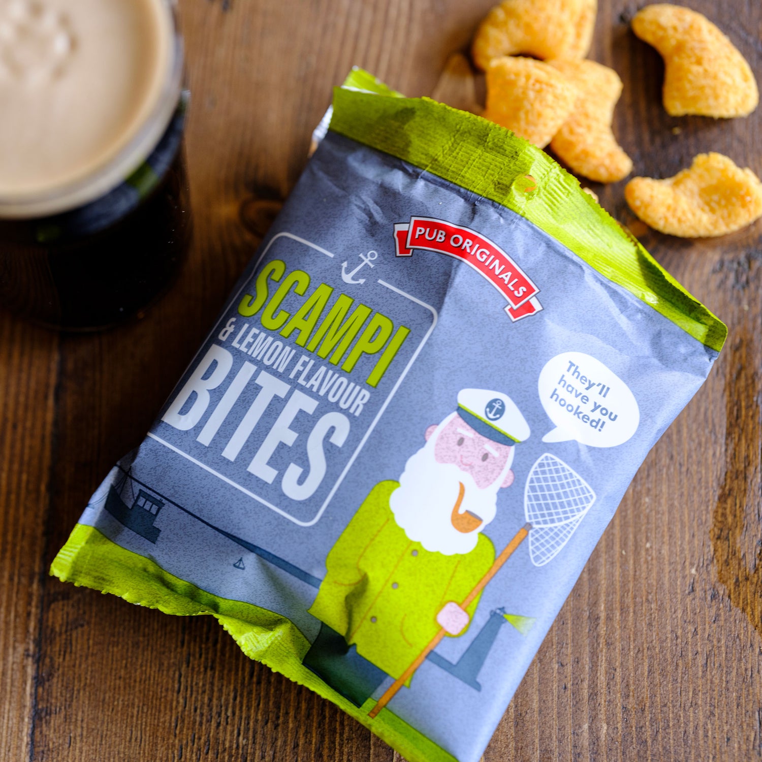 Packaged Pub Originals Scampi & Lemon Flavour Bites on a wooden surface with a stout and snacks.