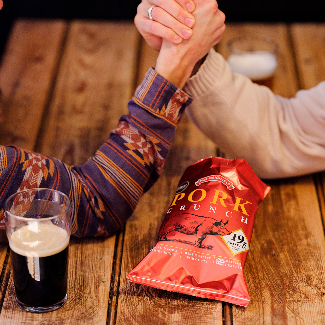 Two people arm wrestling over a packet of Pub Originals Pork Crunch and a pint of beer.